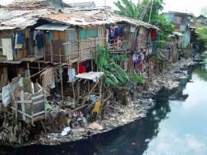 wooden slums in Jakarta stand along the side of a river polluted with trash.