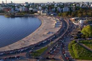 A wide shot from above Montevideo shows buildings along the skyline among trees, a beach next to a bay on the left populated with people enjoying sunset, a park full of picnicers on the right, and busy streets full of cards between the beach and the park.