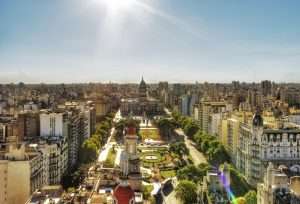 A bright sun shines down on the Palacio del Congreso, in Buenos Aires, Argentina, full of modern and traditional buildings, greenery, and people.